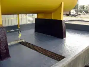 Piso de dique de contenção com revestimento epóxi cinza, suportes de tanque revestidos e grelha de drenagem ao fundo.
