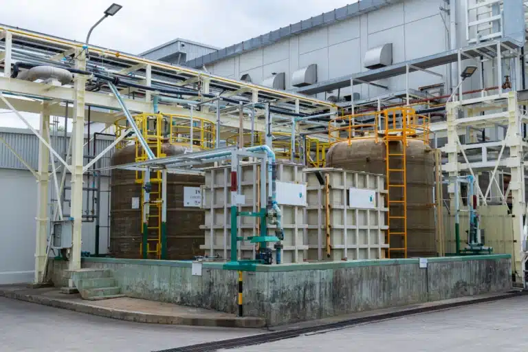 Tanques cilíndricos e modulares de armazenamento químico instalados dentro de bacia de contenção em concreto, com tubulações industriais e estruturas metálicas.