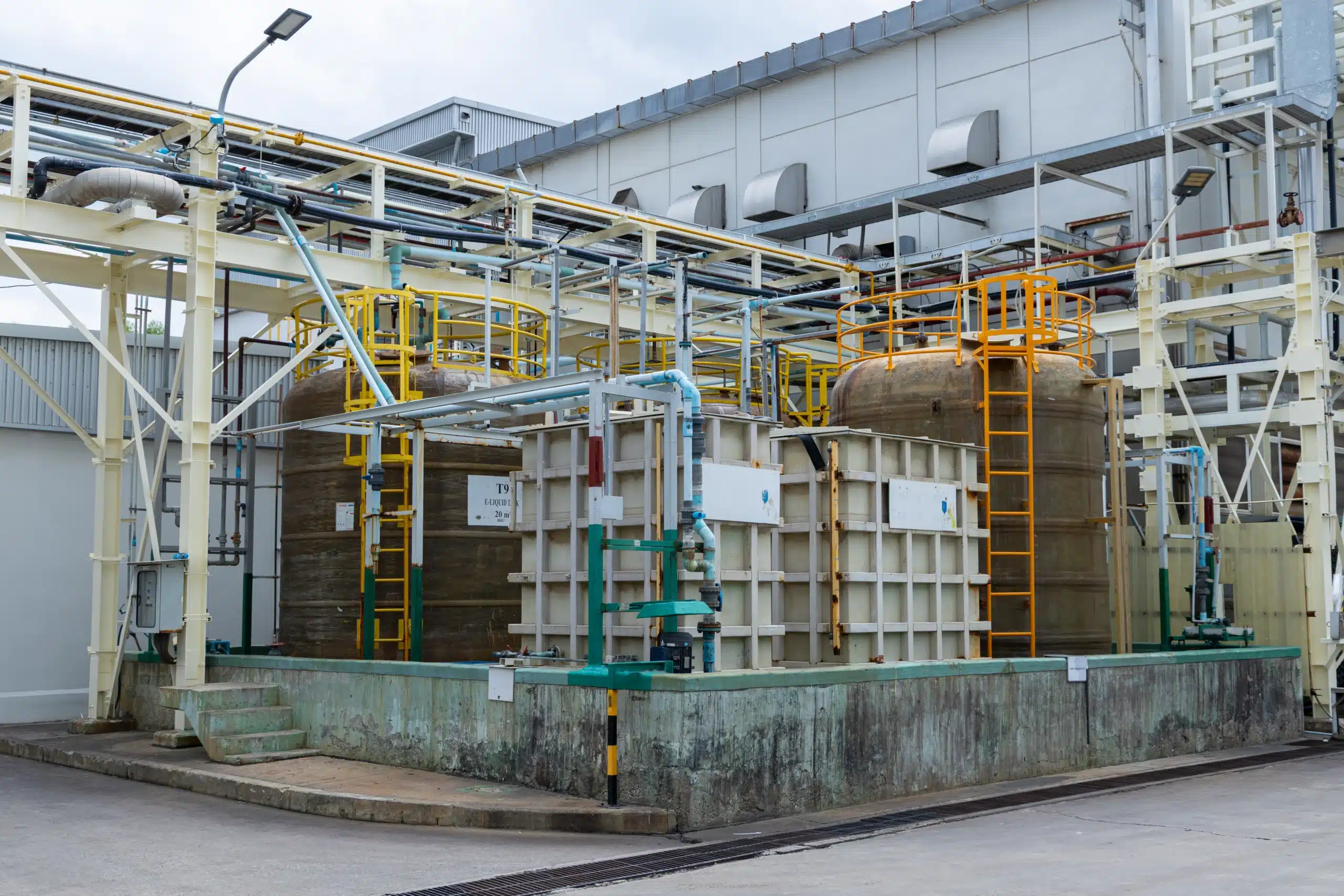 Tanques cilíndricos e modulares de armazenamento químico instalados dentro de bacia de contenção em concreto, com tubulações industriais e estruturas metálicas.