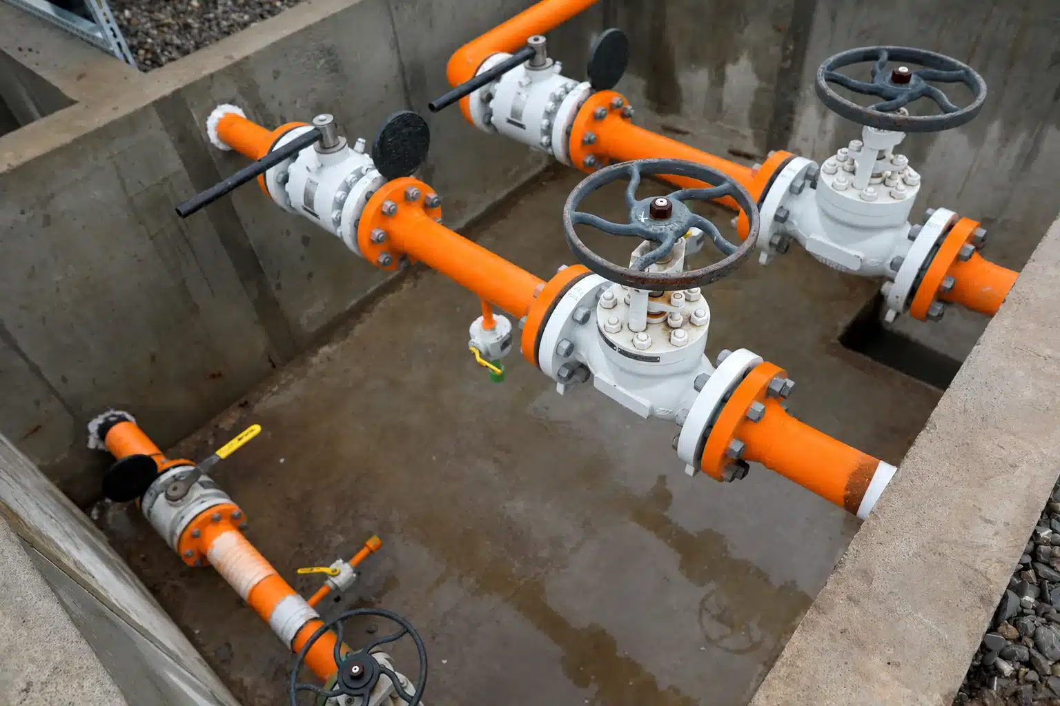 Poço de contenção em concreto contendo tubulações industriais laranjas e válvulas flangeadas com sinais de acúmulo de efluente no piso.